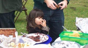Child licking her fingers in front of a table of food at a picnic